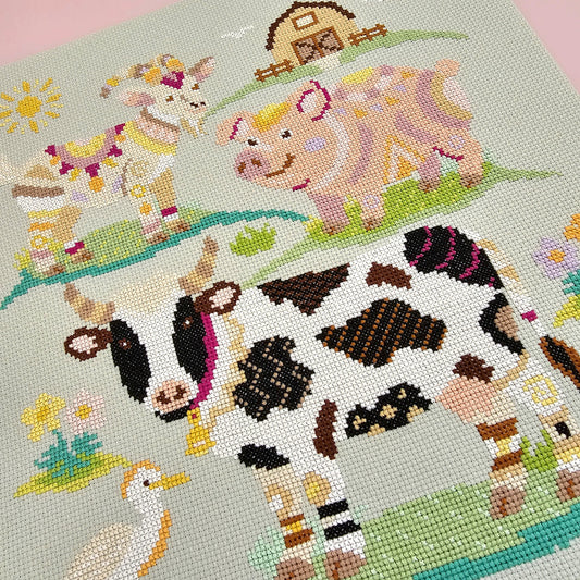 Finished Farmyard Finds cross stitch design flatlay featuring a colourful geometric cow, pig and goat on green aida fabric